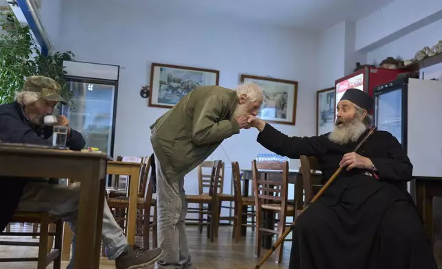 Emanouil IoaNnou Nomikos, 65, kisses the hand of Orthodox Christian monk Father Spyridon of Amorgos as he enters a coffee shop while Evagelos Zak, 70, drinks his coffee at Tholaria village , Amorgos Island, Greece, on Saturday, March 29, 2025. (AP Photo/Petros Giannakouris)