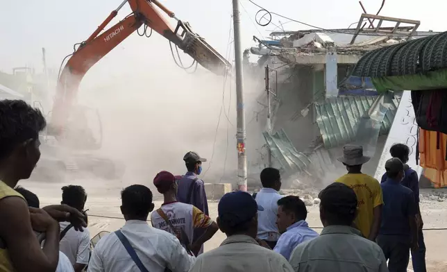 People watch as a backhoe clear debris from damaged buildings at a market, after one month of the strong earthquake in Naypyitaw, Myanmar, Monday, April 28, 2025. (AP Photo/Aung Shine Oo)