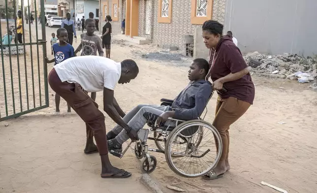 A man helps Adiaratou Ba, right, lift her son Mamadou, 13, suffering from a rare genetic disease, onto the curb in Dakar, Senegal, Saturday Jan. 11, 2025. (AP Photo/Annika Hammerschlag)