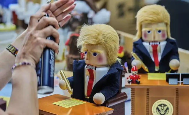 A woman uses hairspray to fixe the hair on a nutcracker titled "Resolute Desk of the President," featuring a seated figure signing a "presidential proclamation," at the Steinbach-Volkskunst nutcracker factory in Marienberg, Germany, Tuesday, April 15, 2025. (AP Photo/Markus Schreiber)