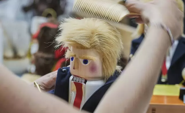 A woman combs the hair on a nutcracker titled "Resolute Desk of the President," featuring a seated figure signing a "presidential proclamation," at the Steinbach-Volkskunst nutcracker factory in Marienberg, Germany, Tuesday, April 15, 2025. (AP Photo/Markus Schreiber)