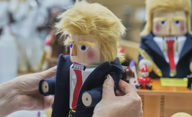 A woman fixes a coat on a nutcracker titled "Resolute Desk of the President," featuring a seated figure signing a "presidential proclamation," at the Steinbach-Volkskunst nutcracker factory in Marienberg, Germany, Tuesday, April 15, 2025. (AP Photo/Markus Schreiber)