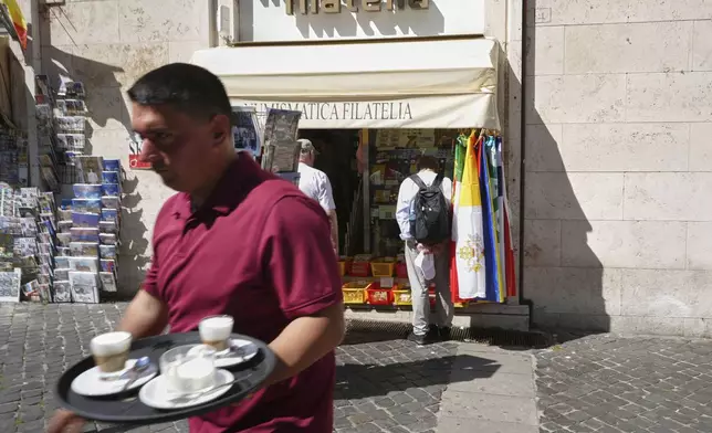 A waiter carries coffees as he walks past the Filitalia stamps, coins, and souvenirs shop just next to the Vatican walls, in Rome, Tuesday, April 29, 2025. (AP Photo/Andrew Medichini)