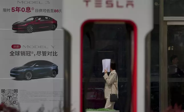 A woman covers her face with a paper as she walks by an electric vehicle charging station outside of a Tesla dealership, in Beijing, Wednesday, April 9, 2025. (AP Photo/Andy Wong)