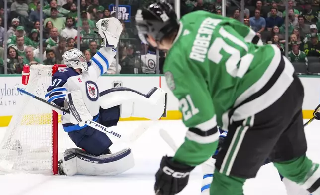 Winnipeg Jets goaltender Connor Hellebuyck, left, catches a shot by Dallas Stars left wing Jason Robertson (21) during the first period of an NHL hockey game Thursday, April 10, 2025, in Dallas. (AP Photo/Julio Cortez)