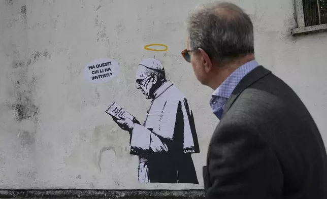 A man walks past a graffiti by artist Laika depicting Pope Francis holding a list of guests for his funeral and reading in Italian "Who invited them?" in Rome, Friday April 25, 2025. (AP Photo/Luca Bruno)
