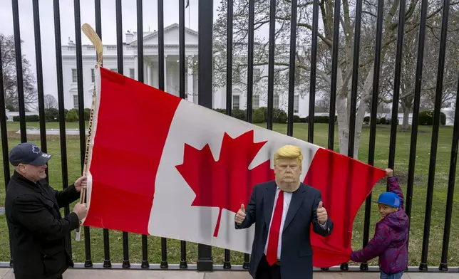 FILE - Toronto residents Douglas Bloomfield, left, and his son Phoenix, right, hold a Canadian flag and an ice hockey stick to show their support for Canada regarding trade tariffs as they pose with with another visitor to the city wearing a mask of President Donald Trump in front of the White House in Washington, March 13, 2025. (AP Photo/Ben Curtis, File)