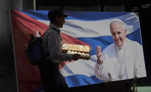 An off-duty police officer carries a carton of eggs past a photo of Pope Francis at the entrance to the car museum where the three Popemobiles he used during his visit to Cuba are parked in Havana, Tuesday, April 22, 2025. (AP Photo/Ramon Espinosa)