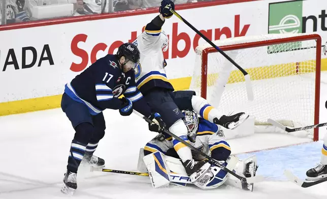 St. Louis Blues' Jake Neighbours, center, falls over Blues goaltender Jordan Binnington, right, as he is checked by Winnipeg Jets' Adam Lowry (17) during the third period of Game 2 of a first-round NHL hockey playoff series in Winnipeg, Manitoba, Monday April 21, 2025. (Fred Greenslade/The Canadian Press via AP)