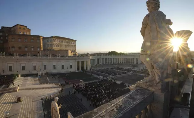 The sun rises through a statue as people begin to take their seats in St. Peter's Square, ahead of the funeral of Pope Francis at the Vatican, Saturday, April 26, 2025. (AP Photo/Markus Schreiber)