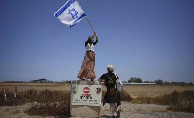 An activist stands on a concrete block signaling border ahead as she waves an Israeli flag towards trucks loaded with humanitarian aid for the Gaza Strip in southern Israel, Wednesday, May 21, 2025. (AP Photo/Ohad Zwigenberg)