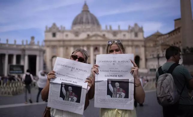 Mackenzie Magas and Kaylee Mellentine from Missouri, pose for a photo with a newspaper showing the new Pope Leo XIV on the front page, in front of the St. Peter Basilica at the Vatican, Friday, May 9, 2025. (AP Photo/Markus Schreiber)