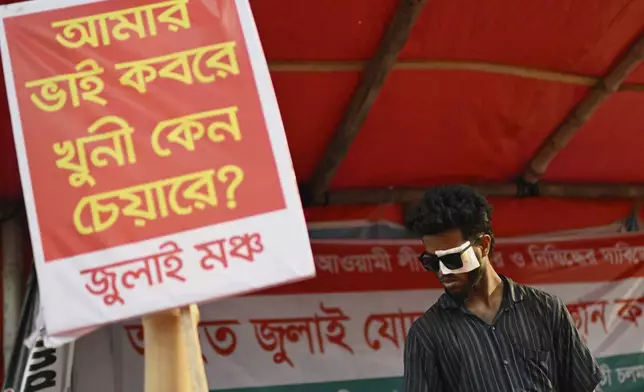 A man who was injured in protests against former Prime Minister Sheikh Hasina last year, stands next to a banner that reads 'My brother is in the grave, why is the murderer in the chair?' during a protest to demand a ban on her Awami League party, in Dhaka, Bangladesh, Sunday, May 11, 2025. (AP Photo/Mahmud Hossain Opu)
