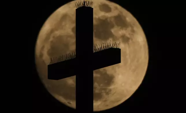 A cross atop the Cathedral of the Immaculate Conception Catholic Church is silhouetted against the rising full moon Sunday, May 11, 2025, in Kansas City, Mo. (AP Photo/Charlie Riedel)