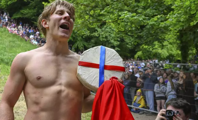 Competitor Tom Kopke of Munich, Germany, holds the cheese after wiining the men's downhill race category of the traditional annual Cheese Rolling contest at Cooper's Hill in Brockworth, Gloucestershire, England, Monday, May 26, 2025.(AP Photo/Anthony Upton)