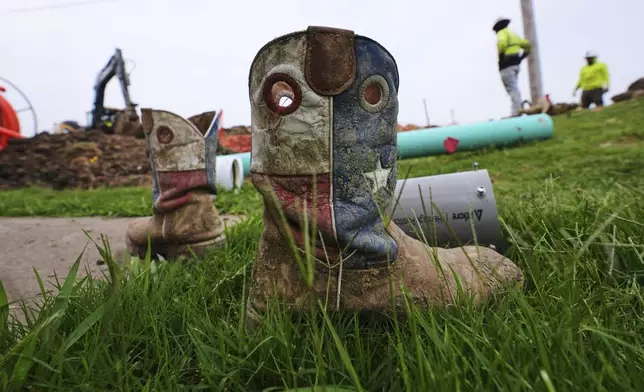 A worker's Texas themed boots sit in the grass as a crew works laying fiber optic cable in Princeton, Texas, Friday, May 16, 2025. (AP Photo/LM Otero)