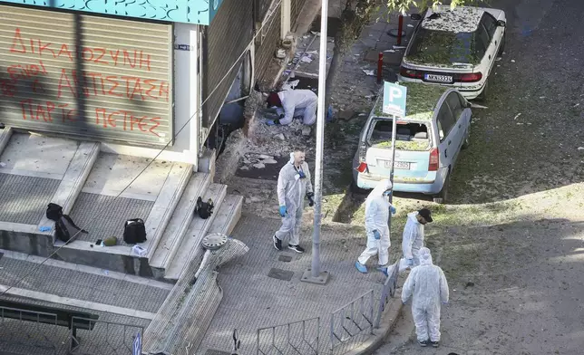 Greek police forensic experts search the area where a 38-year-old woman was killed early Saturday when a bomb she was carrying exploded in her hands outside a bank branch, local police said, in Thessaloniki, northern Greece, Saturday, May 3, 2025. On the fence of the bank branch reads : "Justice is to resist. Take stones". (AP Photo/Giannis Papanikos)
