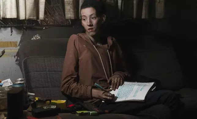A woman looks up from writing in a notebook with the words "to be a princess" seen near the top of the page and a tiara next to her inside a trailer as her father works to get it to start, at a homeless encampment in Deschutes National Forest, Wednesday, April 30, 2025 near Bend, Ore. (AP Photo/Jenny Kane)