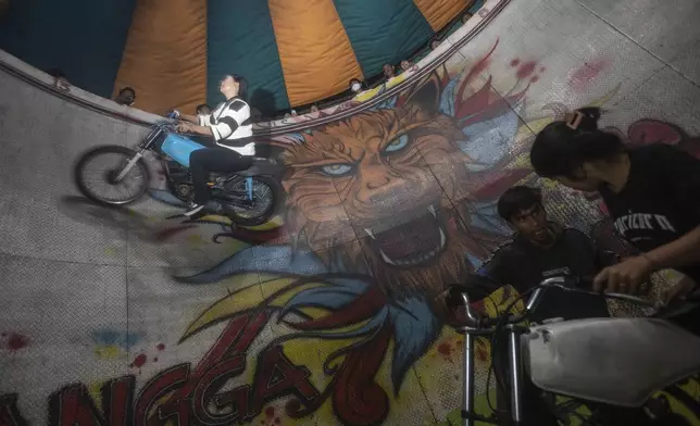 Sri Wahyuni, who is also known as Ayu, top, rides her motorcycle as Rismauly Girsang, bottom right, prepares to perform in the "wall of death," locally known as "tong setan" or satan's barrel, at a fairground in Medan, North Sumatra, Indonesia, Saturday, May 10, 2025. (AP Photo/Binsar Bakkara)