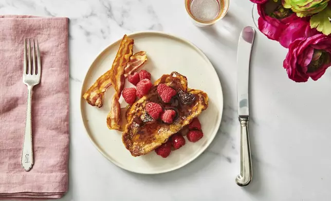 A recipe for challah French toast appears on a plate topped with raspberries, maple syrup, confectioners’ sugar, and a side bacon. (Cheyenne Cohen via AP)