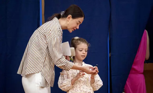 A woman hands a little girl a voting stamp after exiting a voting booth before casting her vote in the first round of the presidential election redo in Bucharest, Romania, Sunday, May 4, 2025. (AP Photo/Andreea Alexandru)