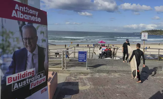 A surfer carries his board as he walks past a picture of Australian Prime Minister Anthony Albanese near a polling booth at Sydney's Bondi Beach, Saturday, May 3, 2025. (AP Photo/Mark Baker)