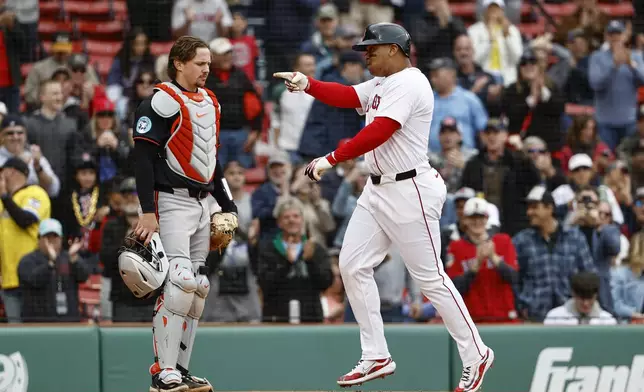 As catcher Adley Rutschman, left, stands at home plate, Boston Red Sox's Rafael Devers, right, points to a teammate as he crosses home after his three-run home run during the sixth inning in the first baseball game of a doubleheader Friday, May 23, 2025, at Fenway Park in Boston. (AP Photo/Winslow Townson)
