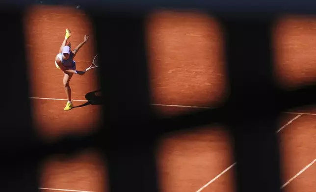 Poland's Iga Swiatek is seen through a fence as she serves against Romania's Jaqueline Cristian during their third round match of the French Tennis Open, at the Roland-Garros stadium, in Paris, Friday, May 30, 2025. (AP Photo/Lindsey Wasson)