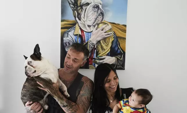 Carolina Morales holds her four-month-old son Benjamin as her husband Alejandro Tirachini holds the family pet Thay, whom they consider their first son, while they pose for a photo backdropped by a painting of Thay by artist Lisandro Guma, in Buenos Aires, Argentina, Friday, April 11, 2025. (AP Photo/Natacha Pisarenko)