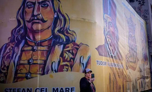A woman holds up a mobile phone backdropped by depictions of Romanian historical figures outside the headquarters of the Alliance for the Unity of Romanians (AUR) shortly before polls close in the first round of the country's presidential election redo in Bucharest, Romania, Sunday, May 4, 2025. (AP Photo/Andreea Alexandru)