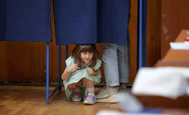 A little girl peers from under a curtain as her father casts his vote in a voting booth in the first round of the presidential election redo in Bucharest, Romania, Sunday, May 4, 2025. (AP Photo/Andreea Alexandru)