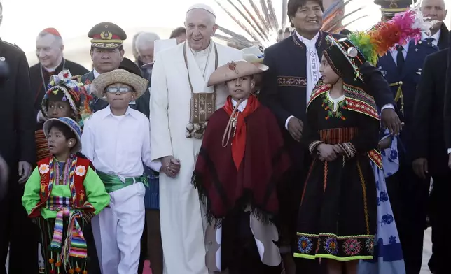 FILE - Pope Francis holds hands with children wearing traditional costumes as he walks with Bolivian President Evo Morales upon his arrival at the airport in El Alto, Bolivia, July 8, 2015. The pouch Francis is wearing around his neck was given to him by Morales. It's woven of alpaca with Indigenous trimmings and traditionally used by people in the Andes to hold coca leaves. (AP Photo/Gregorio Borgia, File)