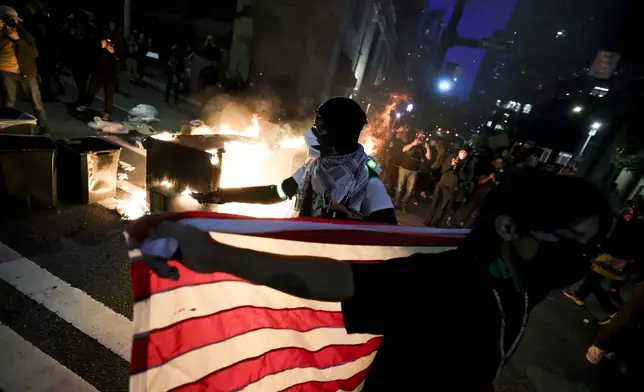A protester holds up an American flag in front of a dumpster that was set on fire during a protest against federal immigration arrests, Wednesday, June 11, 2025, in Seattle. (AP Photo/Ryan Sun)