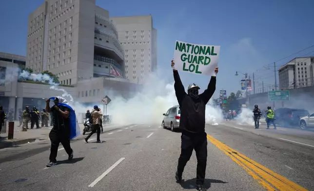 A protester displays a poster as tear gas is used in the metropolitan detention center of downtown Los Angeles, Sunday, June 8, 2025, following last night's immigration raid protest. (AP Photo/Eric Thayer)