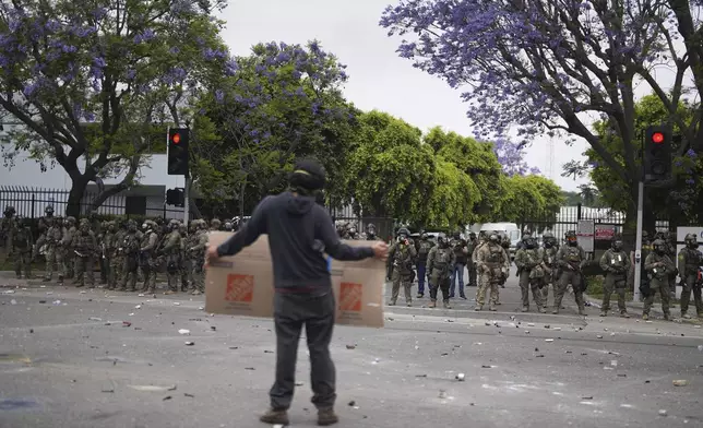 A protester holds a sign as Border Patrol personnel in riot gear and gas masks stand guard outside an industrial park in the Paramount section of Los Angeles on Saturday, June 7, 2025 (AP Photo/Eric Thayer)