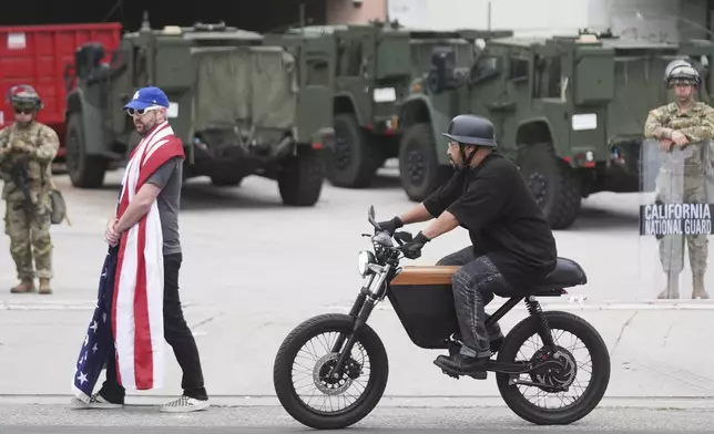 A lone demonstrator draped in a U.S. flag walks outside of the Metropolitan Detention Center Thursday, June 12, 2025, in Los Angeles. (AP Photo/Damian Dovarganes)