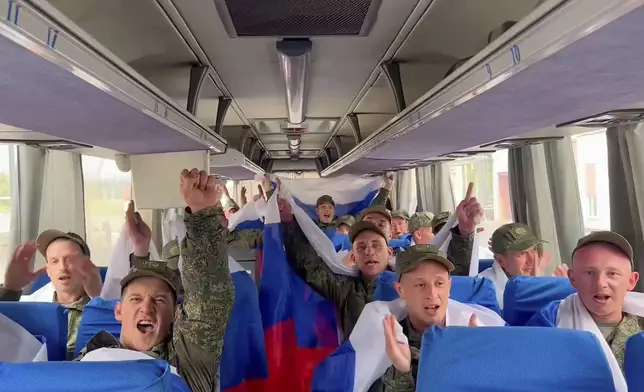 In this photo taken from a video distributed by Russian Defense Ministry Press Service on Friday, June 20, 2025, Russian servicemen wave Russian national flags sitting in a bus at an exchange area in Belarus after returning from captivity during a POWs exchange of a group of servicemen between Russia and Ukraine. (Russian Defense Ministry Press Service via AP)
