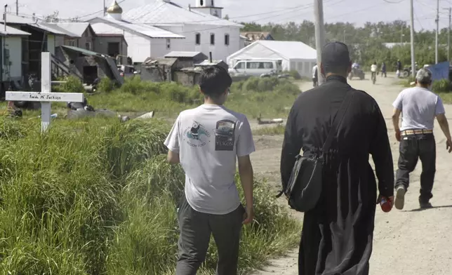 Worshippers, including an Orthodox priest wearing a black cassock, walk on the dusty streets of Kwethluk, Alaska, on June 19, 2025, heading to St. Nicholas Orthodox Church for the canonization ceremony of St. Olga, the first female Orthodox saint in North America. (AP Photo/Mark Thiessen)