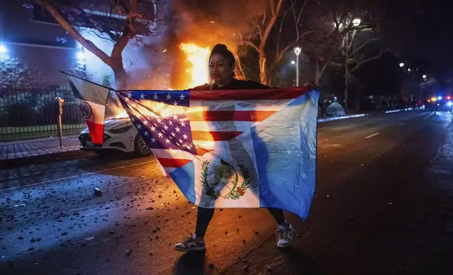 A person displaying multiple flags walks past a burning car during protests over the Trump administration's immigration raids in Los Angeles, Monday, June 9, 2025. (AP Photo/Ethan Swope)