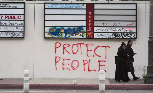People walk past graffiti from recent protests against federal immigration raids, Wednesday, June 11, 2025, in Los Angeles. (AP Photo/Damian Dovarganes)