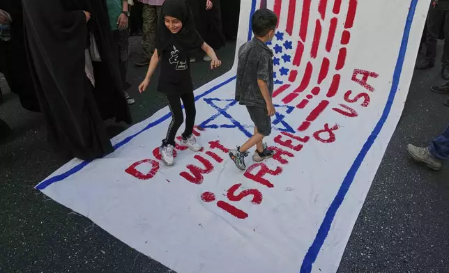Two children walk on an anti-U.S. and anti-Israeli banner during a rally marking the Muslim Shiite holiday of Eid al-Ghadir, which commemorates the Prophet Muhammad naming his successor, Ali, who is revered as the first Shiite imam, in Tehran, Iran, Saturday, June 14, 2025.(AP Photo/Vahid Salemi)