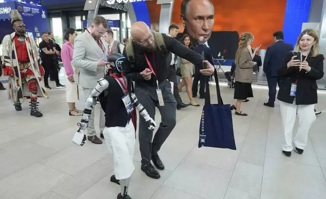 A participants makes a selfie with the Humanoid robot Volodya as other walk past a broadcast of the Russian President Vladimir Putin's speech on a TV screen during a plenary session of the St. Petersburg International Economic Forum in St. Petersburg, Russia, on Friday, June 20, 2025. (AP Photo/Dmitri Lovetsky)