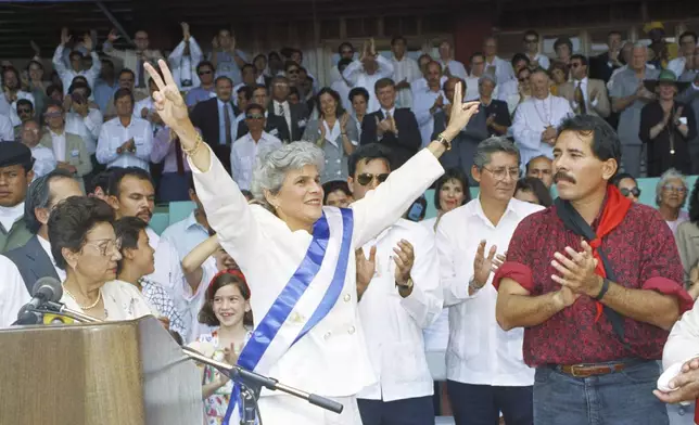 FILE - Nicaragua's new President Violeta Barrios de Chamorro flashes vee signs as outgoing President Daniel Ortega applauds after placing the presidential sash on her, in Managua, April 25, 1990. Barrios de Chamorro, a simple housewife thrust into Nicaraguan politics by the assassination of her husband and who, as president, ended a civil war, died on Saturday, June 14, 2025, in San José, Costa Rica, her family said in a statement. She was 95.(AP Photo/John Hopper, File)