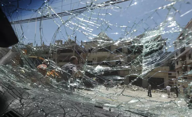 People seen through broken car window, as they gather in front of a destroyed building that was hit in an Israeli airstrike in Nabatieh town, south Lebanon, Friday, June 27, 2025. (AP Photo/Mohammed Zaatari)