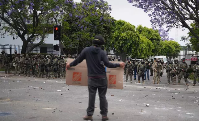 A protester holds a sign as Border Patrol personnel in riot gear and gas masks stand guard outside an industrial park in the Paramount section of Los Angeles on Saturday, June 7, 2025 (AP Photo/Eric Thayer)