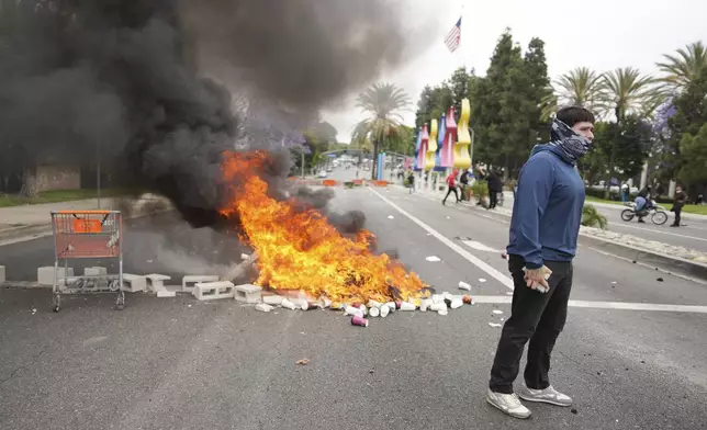 A fire burns as a protester stands across the way from Border Patrol personnel in riot gear and gas masks outside an industrial park in the Paramount section of Los Angeles on Saturday, June 7, 2025 (AP Photo/Eric Thayer)
