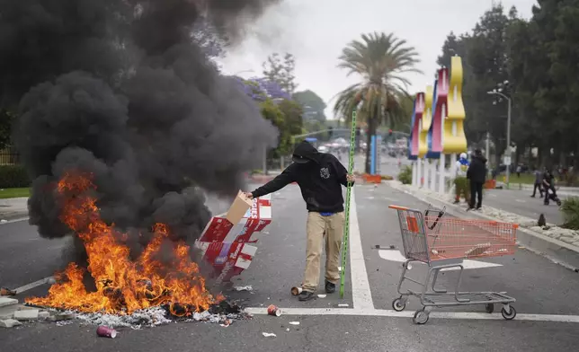 A protester places debris in a fire as Border Patrol personnel in riot gear and gas masks stand guard outside an industrial park in the Paramount section of Los Angeles, on Saturday, June 7, 2025 (AP Photo/Eric Thayer)