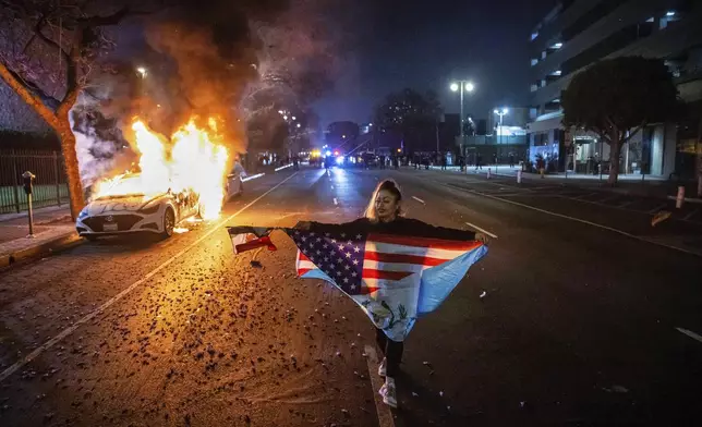 A person carrying multiple flags walks past a burning car during protests over the Trump administration's immigration raids in Los Angeles, Monday, June 9, 2025. (AP Photo/Ethan Swope)