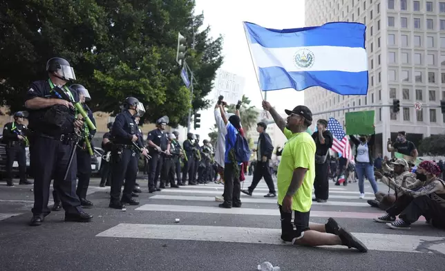 A protester kneels waving a National flag from El Salvador in front of a line of law enforcement officers near a federal building in downtown Los Angeles on Monday, June 9, 2025. (AP Photo/Eric Thayer)
