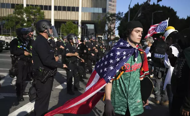 A protester wearing a Los Angeles Dodgers shirt and sporting an American flag as a cape walks past a line of police blocking a street in downtown Los Angeles on Monday, June 9, 2025. (AP Photo Jae Hong)